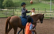 Ein Kind sitzt auf einem Pony, das von einer Reitlehrerin geführt wird und wirf einen Ball zu einem anderen Kind, das nebenher geht.