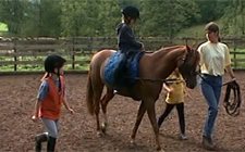 Ein Kind sitzt auf einem Pony, die Reitlehrerin führt das Pony an der Leine, zwei Kinder gehen neben dem reitenden Kind.