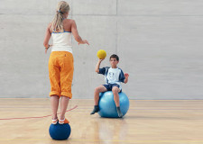 Une fille est debout en équilibre sur un medecine ball et s'apprête à attraper un ballon lancé par un camarade assis sur un ballon de gymnastique.