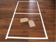 Un rettangolo disegnato a terra con del nastro adesivo bianco, al centro due buste di cartone