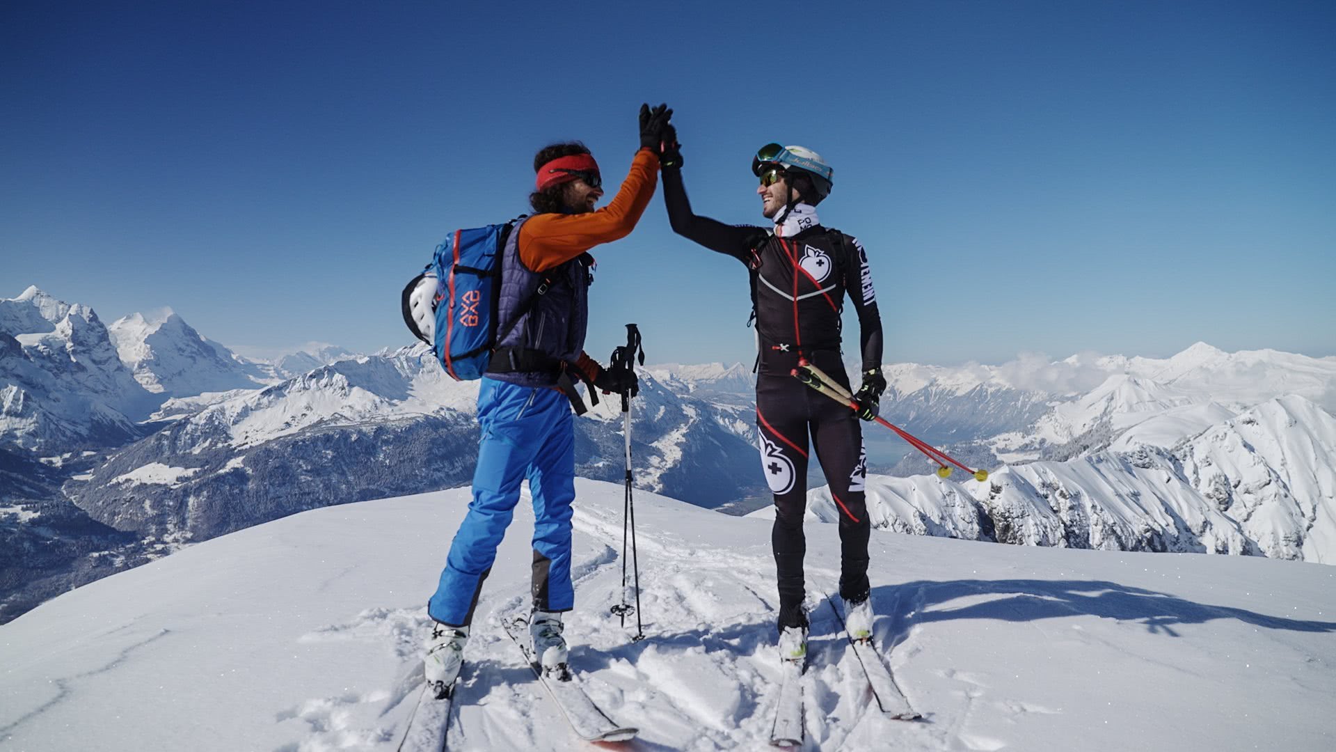Zwei Skitourengänger (klassisch und Wettkampf) klatschen sich auf einer Bergspitze ab.