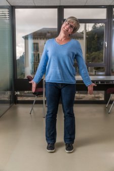 Una donna in piedi in un ufficio esegue un esercizio di allungamento e rilassamento con gambe leggermente divaricate e testa inclinata di lato 