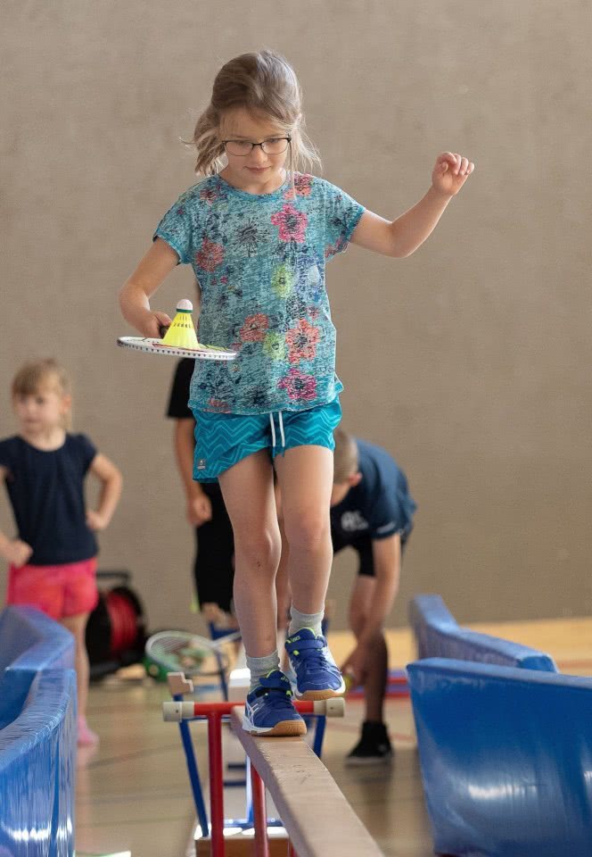 Mädchen balanciert auf schmaler Seite von  Langbank einen Badminton-Schläger mit Shuttle.