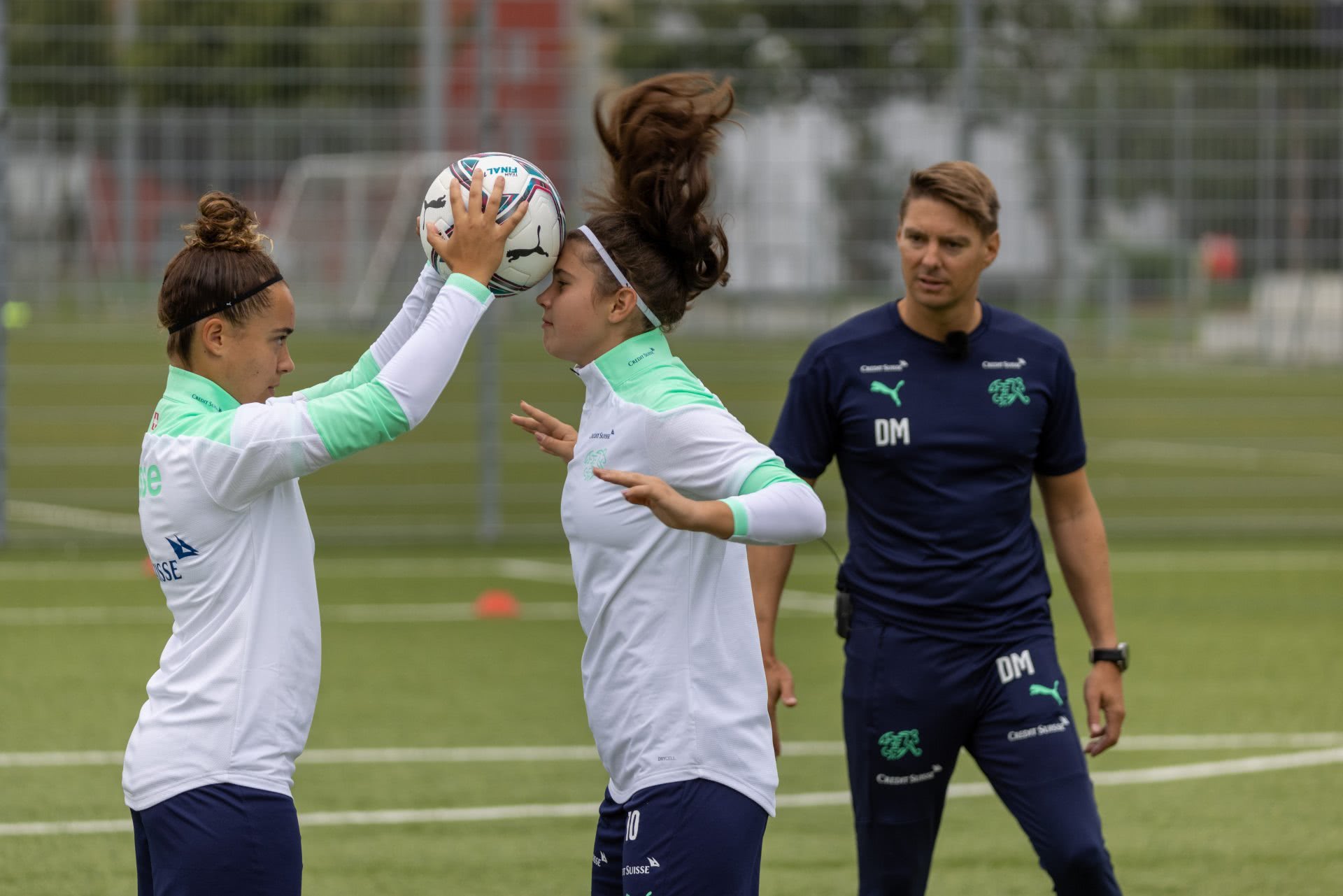 Deux jeunes footballeuses effectuent un exercice du jeu de tête sous le regard de leur entraîneur.