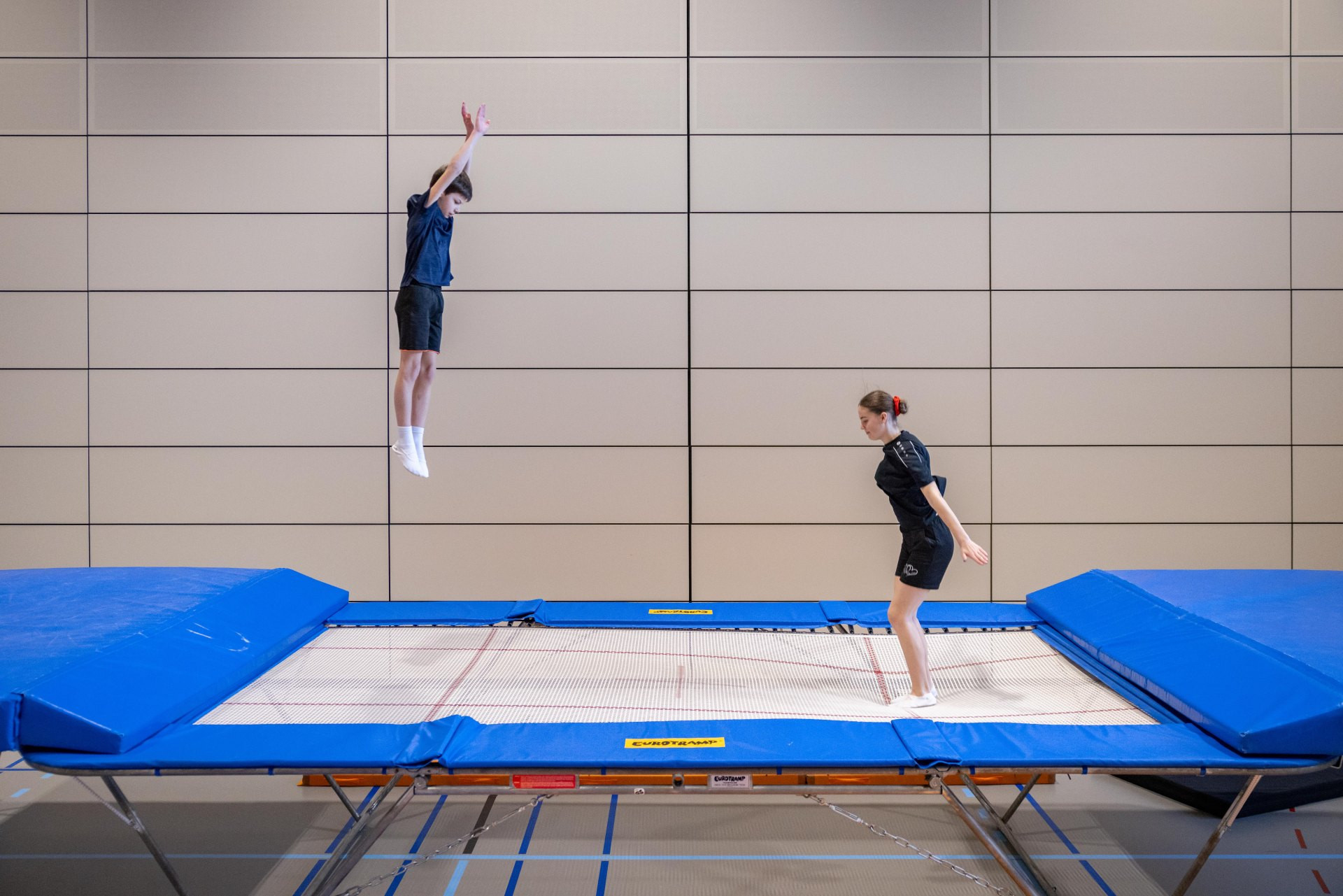 Due ginnasti saltano in modo asincrono sul trampolino. Entrambi eseguono salti in posizione eretta e cercano di raggiungere e mantenere la stessa altezza.
