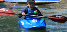 Kind mit Cap sitzt im Kanu und paddelt auf Fotografen zu.