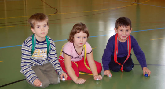 3 Kinder sitzen am boden in einer Reihe unsd spielen mit Flaschendeckeln.