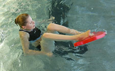 Eine Frau sitzt im Wasser und hält die Füsse auf ein Schwimmbrett. 