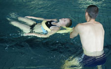 Un uomo trascina una compagna sdraiata sulla schiena in acqua. La donna ha un tubo di gomma sotto le braccia.