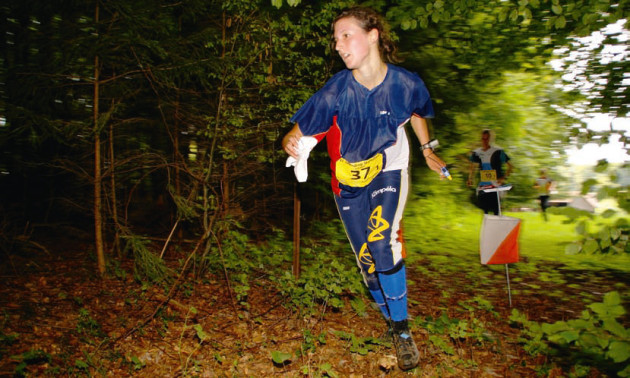 Una giovane corre in un bosco con una cartina in mano seguita da altri due concorrenti