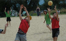 Spiel auf Sand. Ein Kind versucht, einen Ball abzufangen, der über ihn fliegt. Ein anderes fängt den Ball dahinter auf.