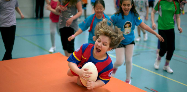 Un bambino con una palla da rugby in mano salta su un tappetone seguito da altri bambini.