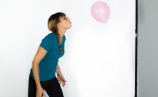 Frau steht vor Wand und bläst einen Luftballon in die Höhe.