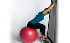 Une femme est assise sur un ballon de gymnastique et tend ses bras au-dessus de sa tête, en appui contre un mur.