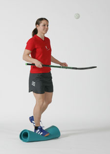 Eine Athletin steht einbeinig auf einer zusammengerollten Gymnastikmatte und jongliert mit Unihockey-Stock und Ball. 