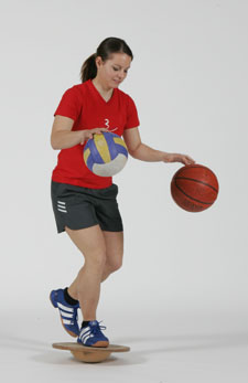 Une athlète est en équilibre sur un ballon et fait rebondir un ballon de basketball et un ballon de volleyball.