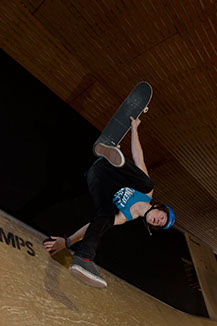 Uno skateboarder solleva i piedi e la tavola appoggiandosi ad una mano su una vertramp.