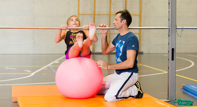 Auf einem Gymball sitzend, schwingt sich eine Schülerin unter einer Reckstange durch. Der Sportlehrer kniet daneben, bereit, im Bedarfsfall einzugreifen. 