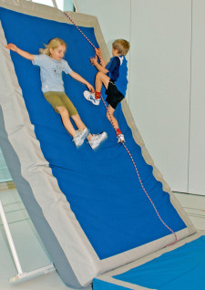 Un enfant monte à une corde attachée aux perches, une autre glisse sur un gros tapis appuyé contre ces mêmes perches.