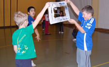 Zwei Kinder halten eine Zeitung mit einem Loch in der Mitte, ein anderes versucht einen Ball hindurch zu werfen.