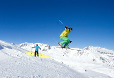 Skifahrer bei einem Sprung, jemand schaut vom Pistenrand zu.