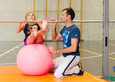 Foto: una bambina è seduta su un pallone da ginnastica e si prepara ad eseguire un movimento difficile assicurata da un insegnante
