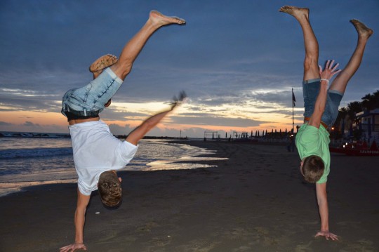 Foto: due giovani mentre eseguono una verticale su un braccio solo su una spiaggia davanti a un bel tramonto