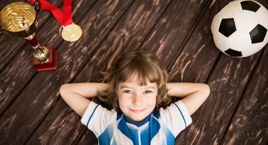 Una bambina sorridente è sdraiata per terra accanto a una coppa, una medaglia e un pallone da calcio