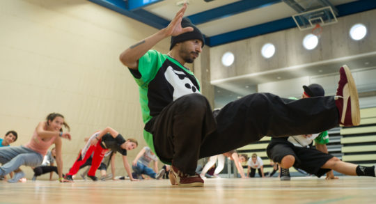 Foto: Lehrperson macht einen Breakdance-Move, Schülerinnen machen ihn nach.