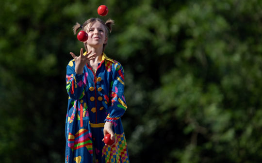 Foto: Mädchen in Clownkostüm beim Jonglieren mit drei roten Bällen.